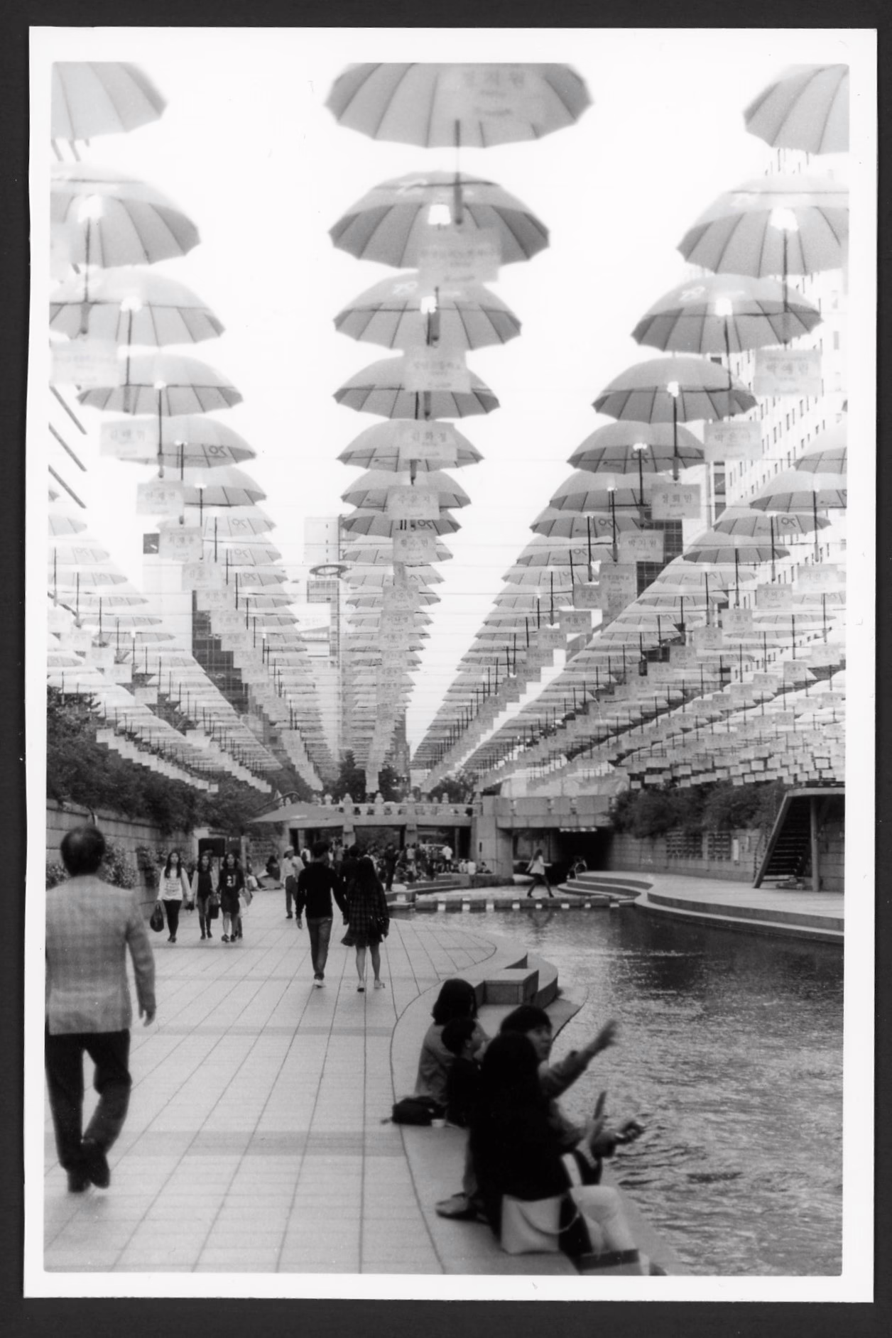 Black-and-white photo Cheonggyecheon stream in Seoul, South Korea. A stream is flanked by two walkways and some small stone bridges cross the river. Dozens of people walk past the stream or sit and chat with each other. Hundreds of umbrellas are strung across the sky above the stream.