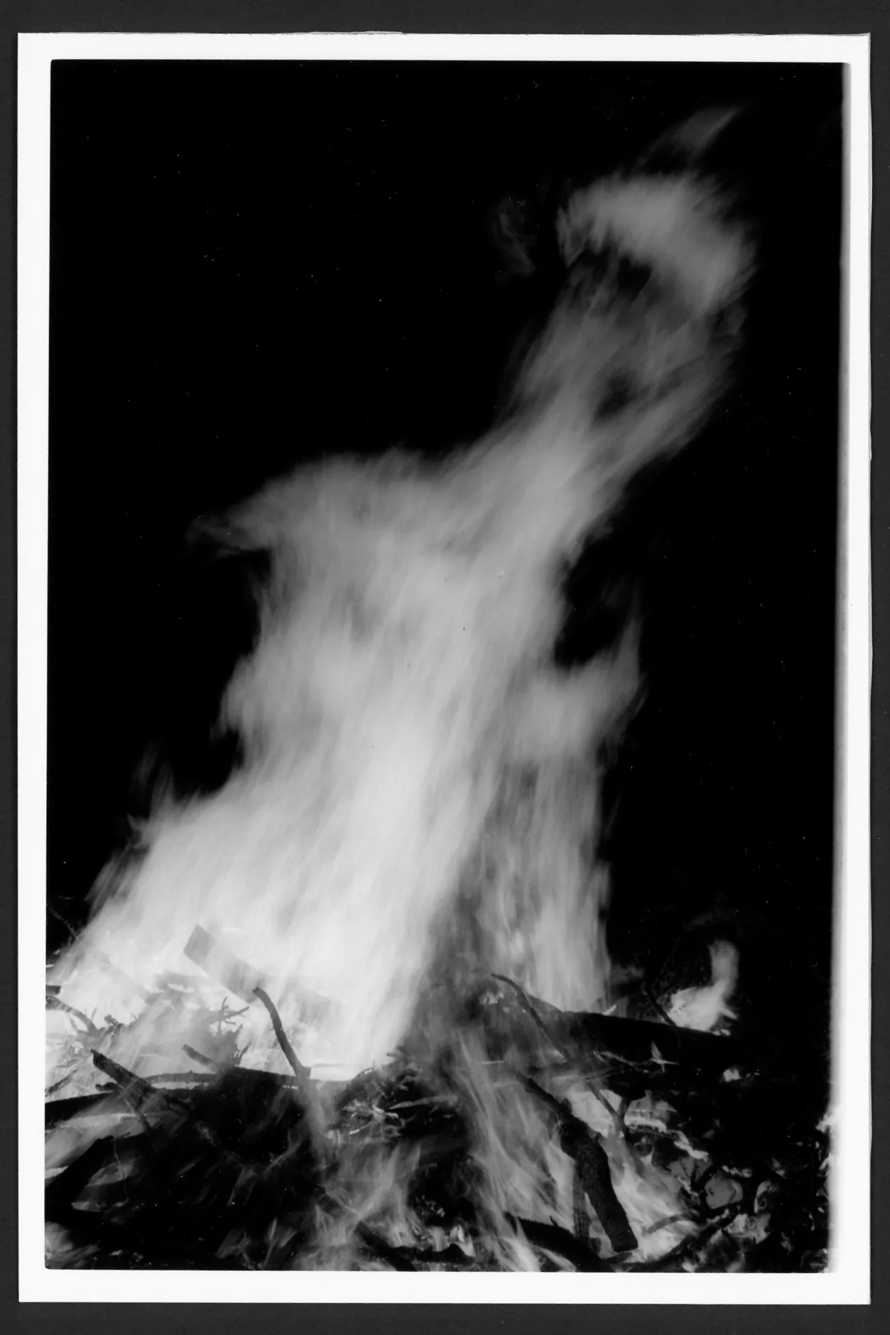 This black and white photo shows a large bonfire. The bottom of the photo shows a mess of sticks on fire, and the fire flames blur together into a mostly smooth surface, albeit one with some texture to it.