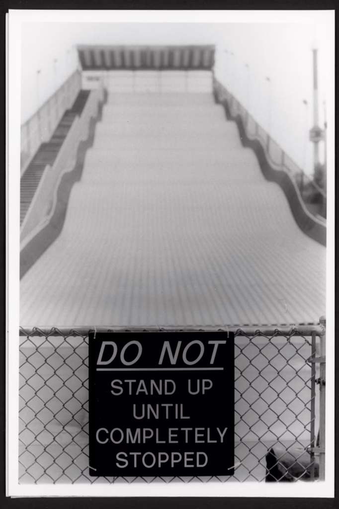 This black and white photo shows the Giant Slide at the Minnesota State Fair fairgrounds. The slide is a closed and a sign attached to a chain link fence says "DO NOT stand up until completely stopped."