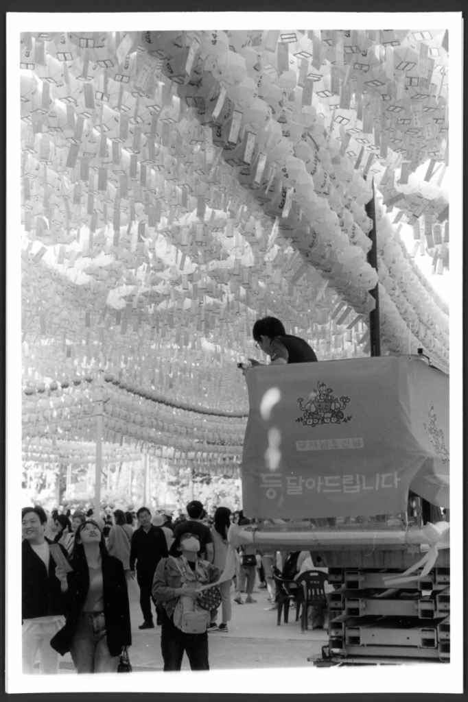 This black and white photo shows a man in a bucket truck hanging lanterns overhead at a Buddhist temple. Below, a crowd of people look up at the lanterns. One woman appears to be asking the man a question.