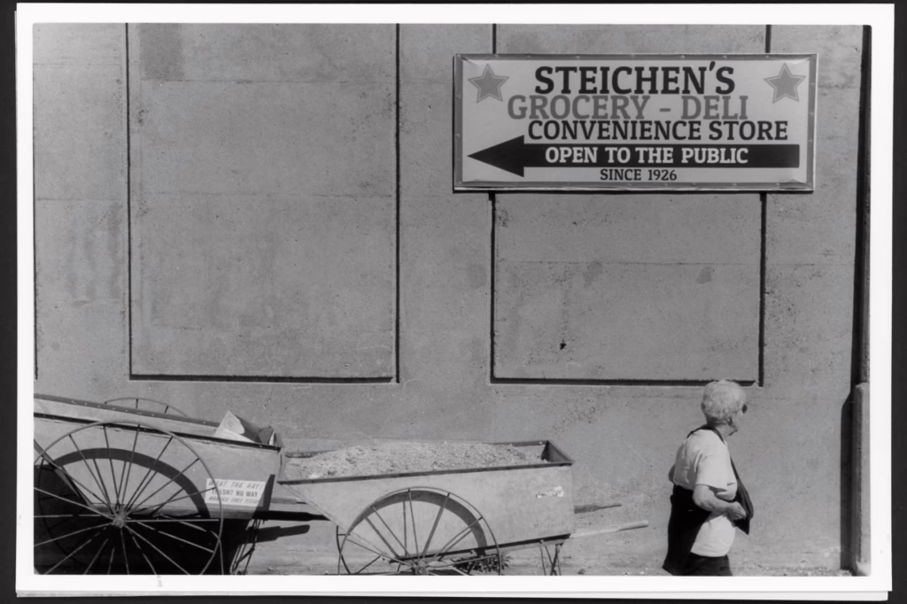 A woman wearing sunglasses walks past a large wall. A sign above her head reads "Steichen's Grocery - Deli Convenience Store Open to the Public Since 1926." Two carts sit in front of the wall. One appears piled high with manure. The other cart has a sticker on it that says "What the Hay! Trash? No way! Manure only today!" That carts appears to have trash in it.