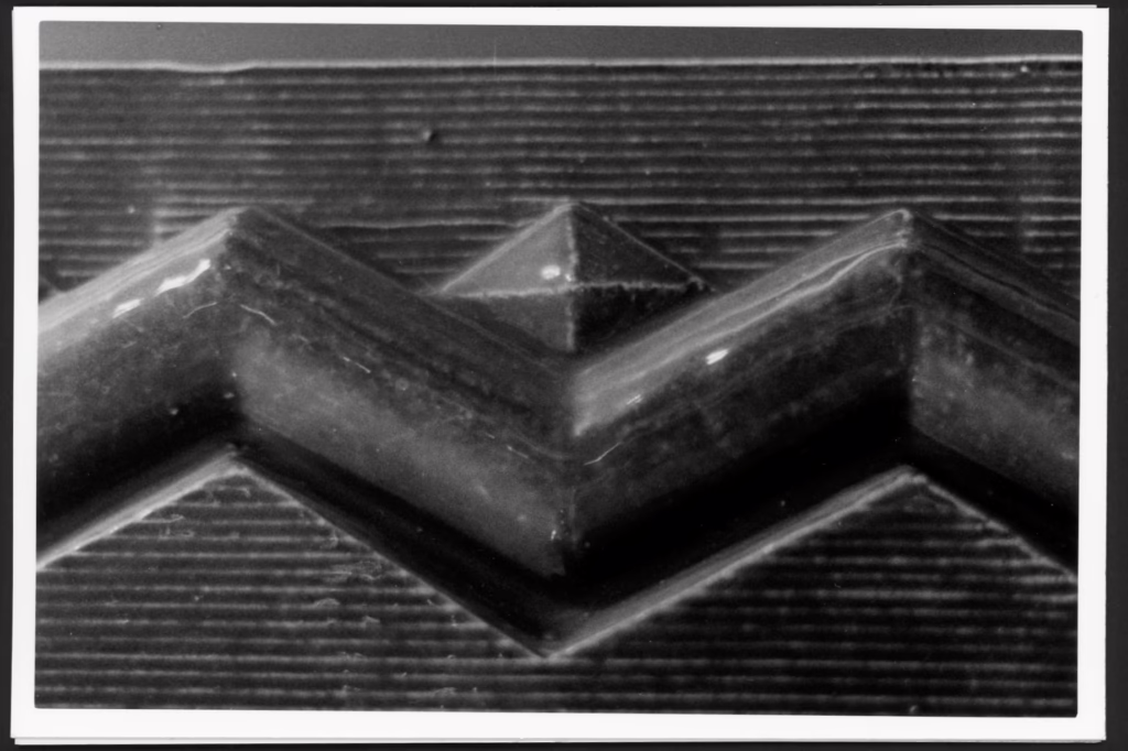 This black and white photo shows a zigzag shape with a diamond on top of it against a thinly striped background. The surface of the zigzag is scratched and chipped.