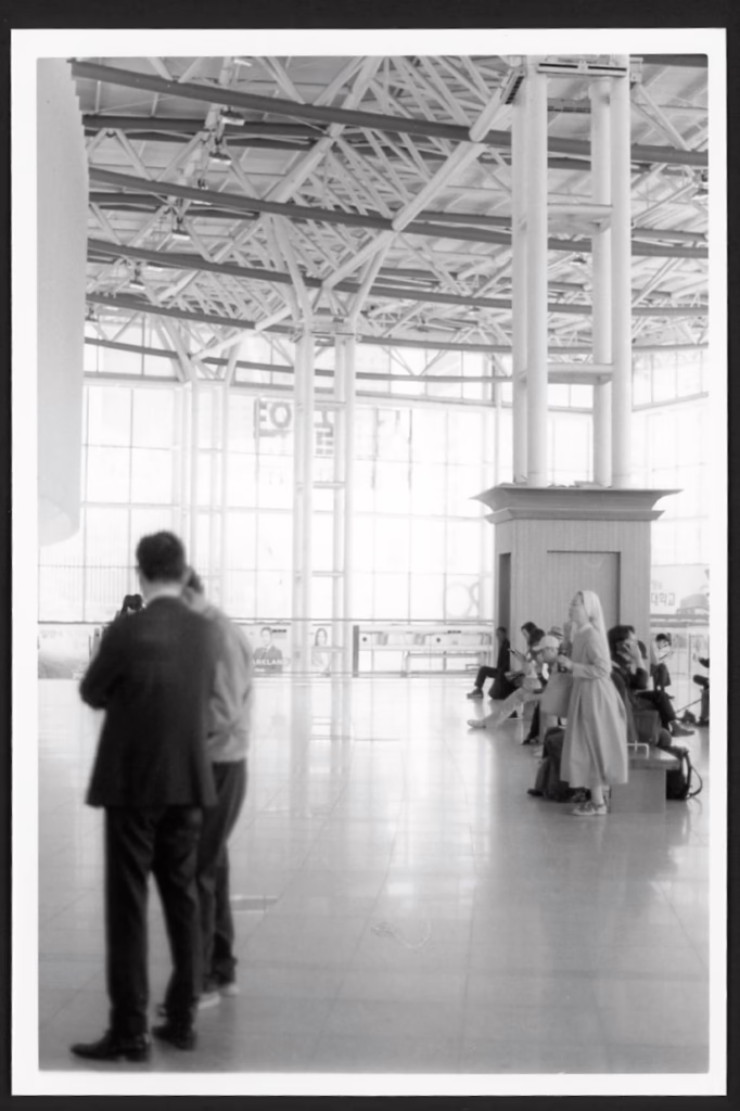 This black and white photograph shows several people waiting at Seoul Station. You can see people standing and seated. Some are in suits, some are in more casual clothing, and one is in a Catholic nun's habit. Sunlight streams through the tall glass walls of the station.