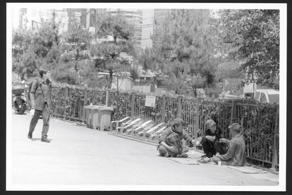 This black and white photo shows three men sitting on the sidewalk in front of a fence in Seoul, South Korea. The three men appear deep in conversation. Another man walks by, looking off into the distance.