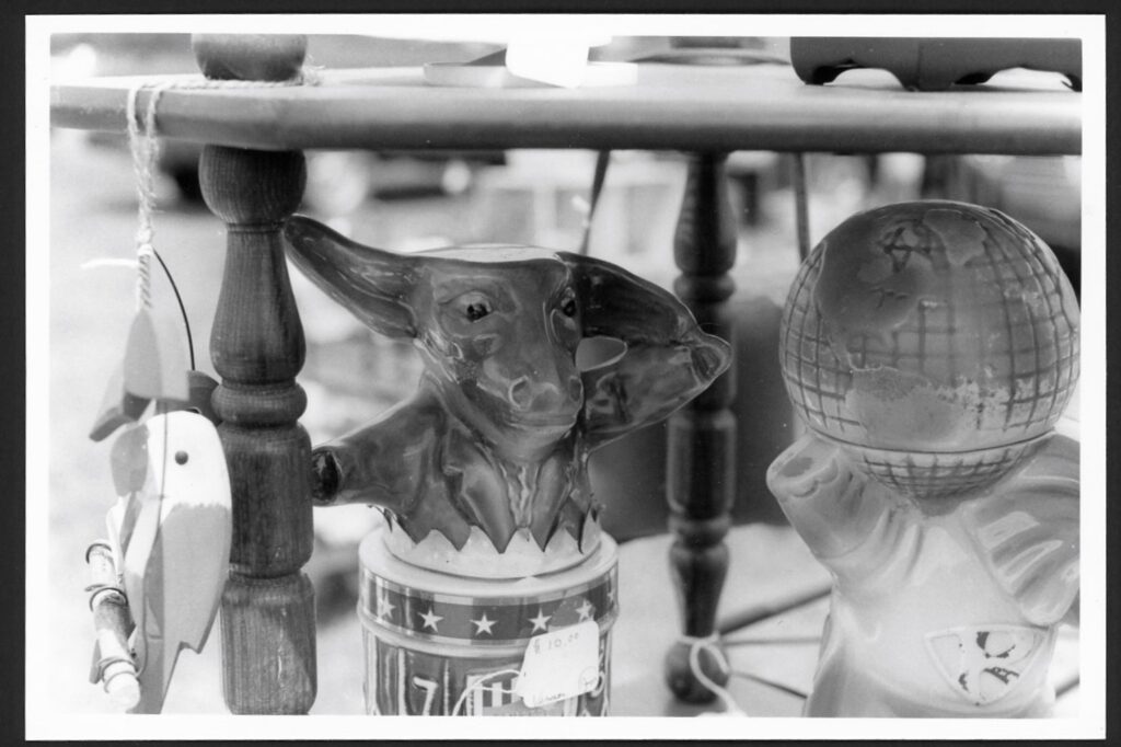 This black and white photograph shows some toys at a flea market. There is a wooden fish, a shiny pigs wearing a tuxedo while popping out of a can, and a globe being held up by an animal of some sort.