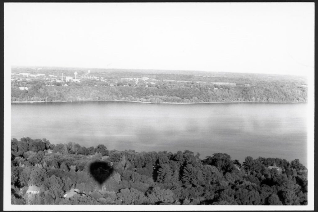 This black and white photograph was taken from a hot air balloon near Stillwater, Minnesota. In the foreground, you can see tree tops, a few roofs, and the balloon's shadow. In the background, you see trees and buildings across the river in Wisconsin. In the midground, there is the St Croix River. The river had an odd texture to it, giving it darker and lighter bubbles of exposure.