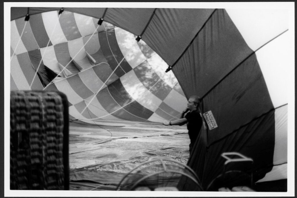 This black and white photograph shows a hot air balloon on its side. The basket and balloon are clearly seen while a large fan can be seen on the bottom edge of the photo. A younger person is helping to fill the balloon.