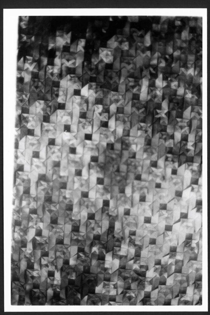 This black and white photograph shows paper folded into origami shapes. The paper alternating dark and light shades, making the image have a repeating pattern of negative space. Only the corner of the photo is in focus, so the photo almost looks like a shattered mirror or perhaps a paper screen of some sort.