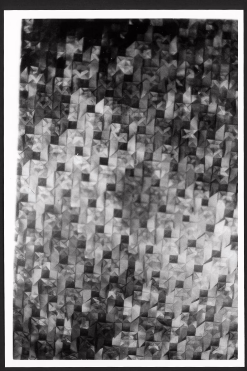 This black and white photograph shows paper folded into origami shapes. The paper alternating dark and light shades, making the image have a repeating pattern of negative space. Only the corner of the photo is in focus, so the photo almost looks like a shattered mirror or perhaps a paper screen of some sort.