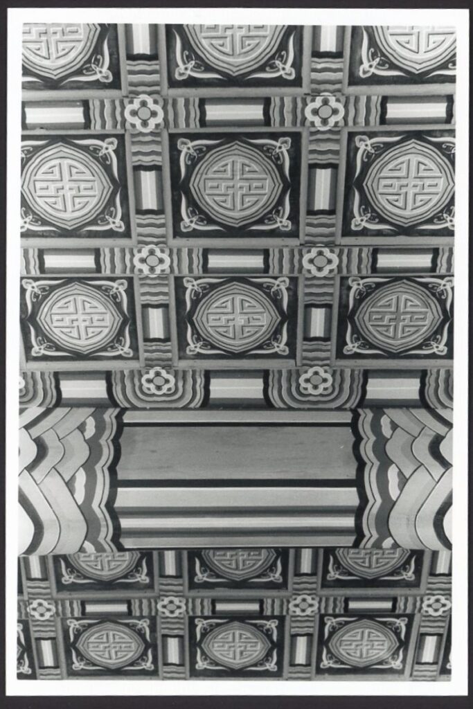 This black and white photo shows a patterned ceiling in a temple in Seoul South Korea. The primary medallion is a large circle with an angular motif in the middle. Between each medallion is a simple floral pattern.