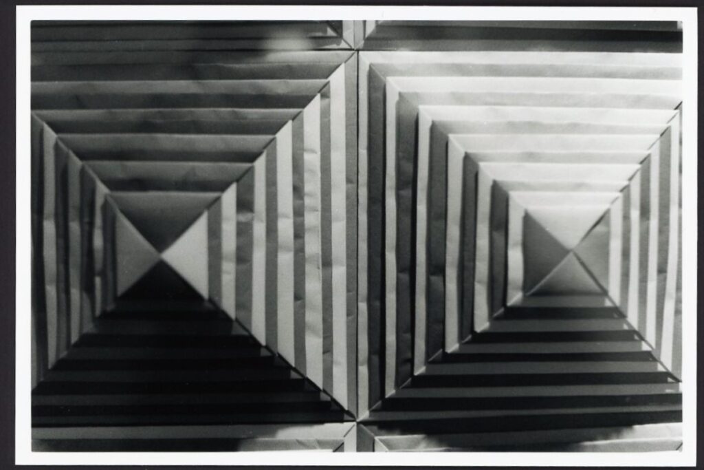 This black and white photo shows pyramids made out of paper. These pyramids are on a vertical surface so the points stretch out from the canvas. The pyramids are made with different colors of paper, making stripes on the shapes.