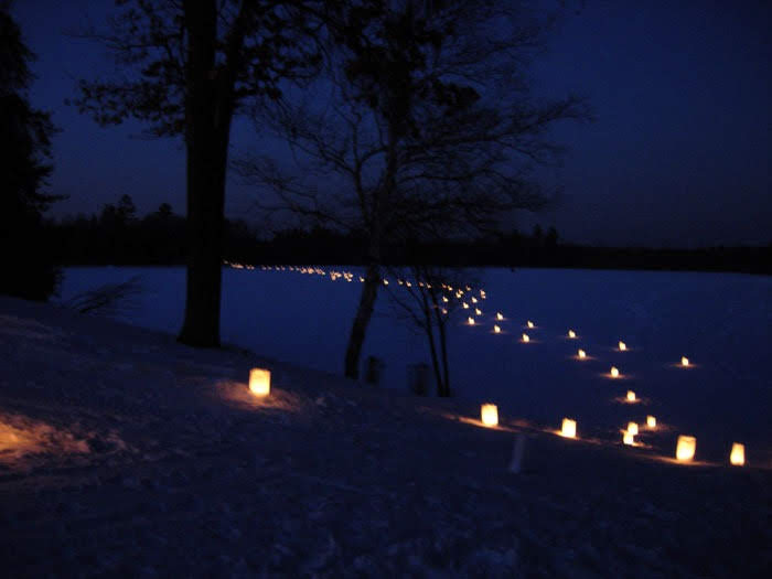 This photo shows a series of candles along a snowy path in the evening. The sky and snow are a dark blue and trees are seen in the foreground and distance.
