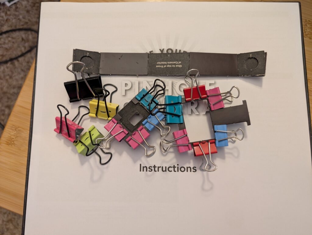 This color photo shows a pile of small binder clips. Some of the binder clips are being used to hold pieces of cardboard together as glue dries. These all rest on some instructions for building a pinhole camera from a kit.