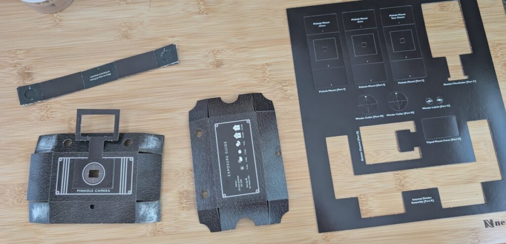 This photo shows various parts of tagboard being used to create a pinhole camera from a kit. You can see how parts get punched out of paper, glued, and held together with binder clips.