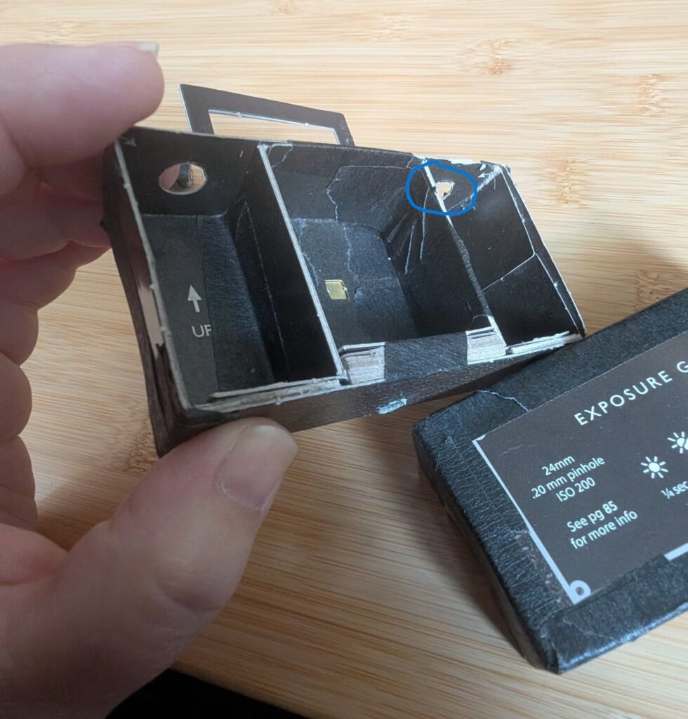 A tagboard pinhole camera is in two parts. Someone is holding one half of the camera. A blue circle points to a tiny point of plastic that is used as an auditory clicker.