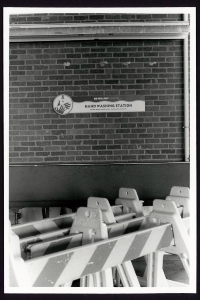 Traffic barricades are stacked one behind the other in front of a brick wall. A sign reading "Be Healthy & Fair: Handwashing Station" can be seen in the background.
