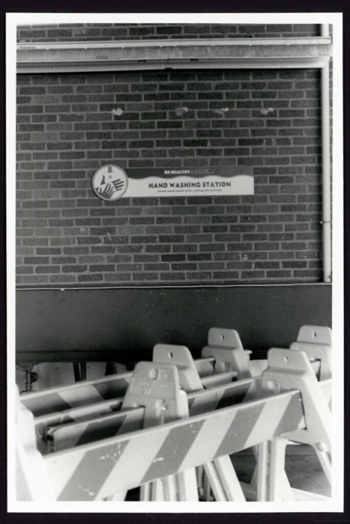 Traffic barricades are stacked one behind the other in front of a brick wall. A sign reading "Be Healthy & Fair: Handwashing Station" can be seen in the background.