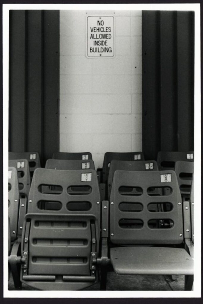 This black and white photo shows stacks of attached folding chairs, like those found in a stadium, stacked up in three rows. On the wall behind the chairs, a sign reads "No Vehicles Allowed Inside the Building."