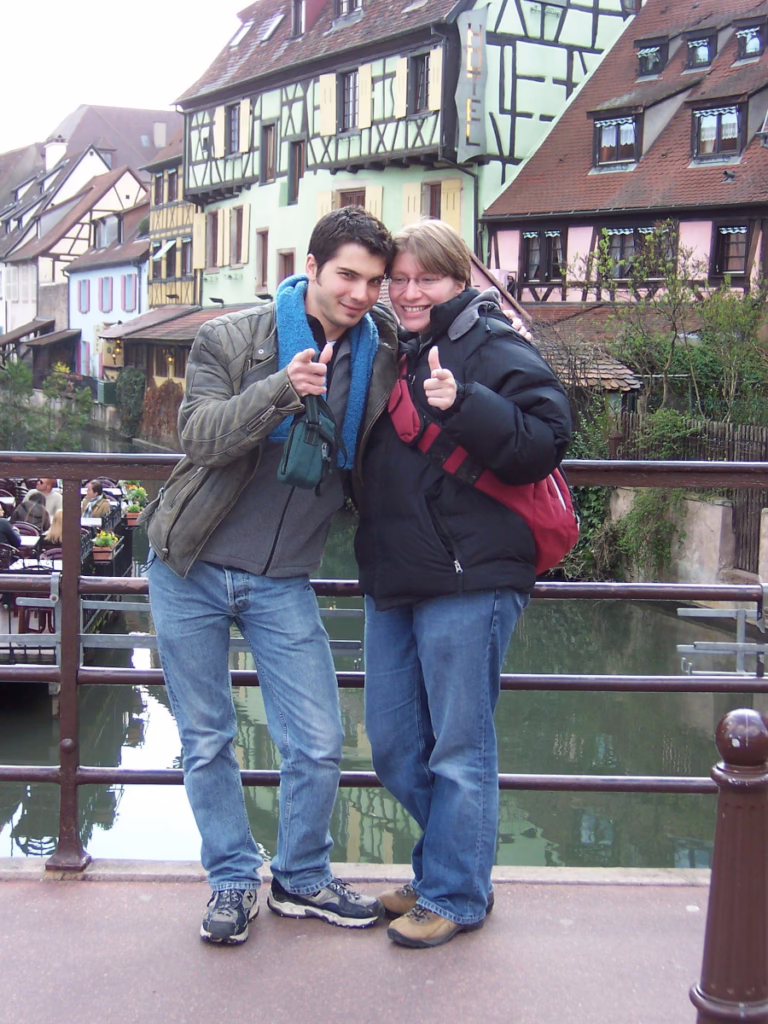 Two people stand in front of some water in Colmar, France, 2006. A very picturesque village can be seen in the background.