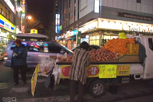A middle-aged Korean woman stands in front of a flat bed truck covered in apples and oranges. A man stands nearby. Cars and other shops can be seen in the background.