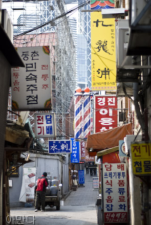 A narrow street sits between tall buildings. Billboards and advertisements in Korean and traditional Chinese characters can be seen. Someone pushes a car in the midground.
