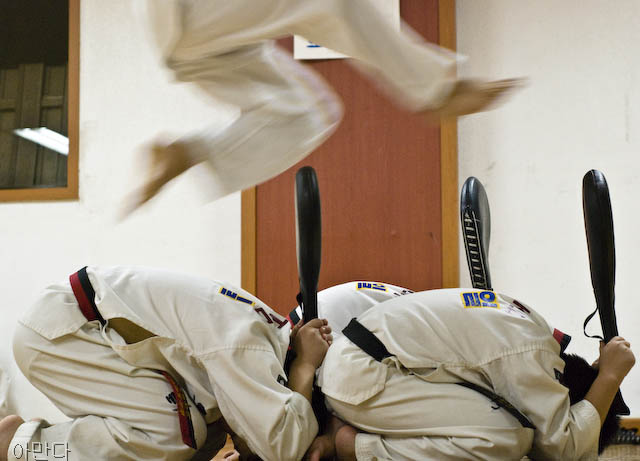 Three taekwodo students sit on the floor on their knees, heads curled into their knees so their bodies form tight balls. Each student holds a kicking target. A fourth student leaps above their bodies, his feet ready to kick each board.