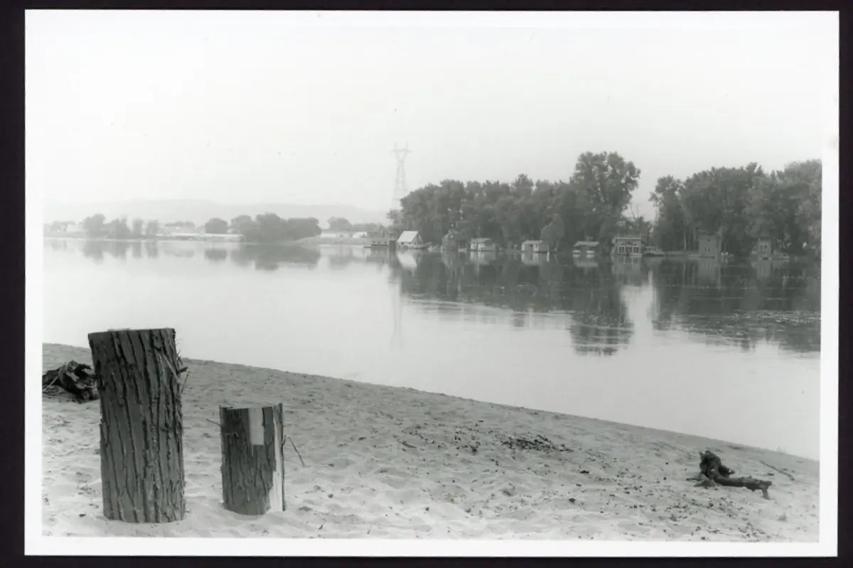This black and white photo shows a sandy beach along the edge of a river. There are some burnt logs on the beach and on the other side of the river you can see a string of boathouses.