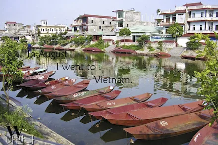 A photo shows boats on a river in Vietnam. Across the river, buildings can be seen. The sky is a pale blue and green plants flank the edges of the photo. The letter W is in the lower corner of the photo.