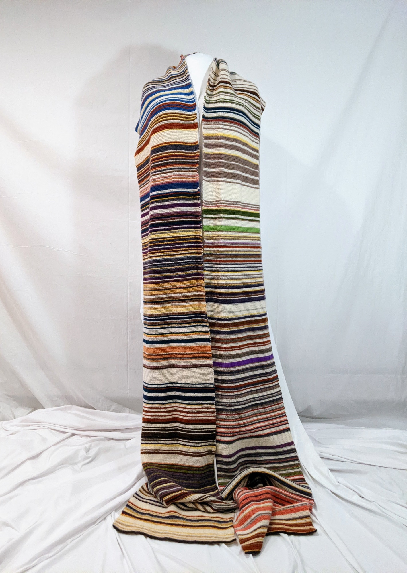 A very long striped scarf is arranged on a dress form. This scarf uses color coding to show the daily work I did in my doctoral program, from the day I passed my oral exams to the day of my dissertation defense. Shades of purple are writing tasks, shades of yellow are administrative work, white is rest, and that odd pearl grey color shows my cancer appointments.