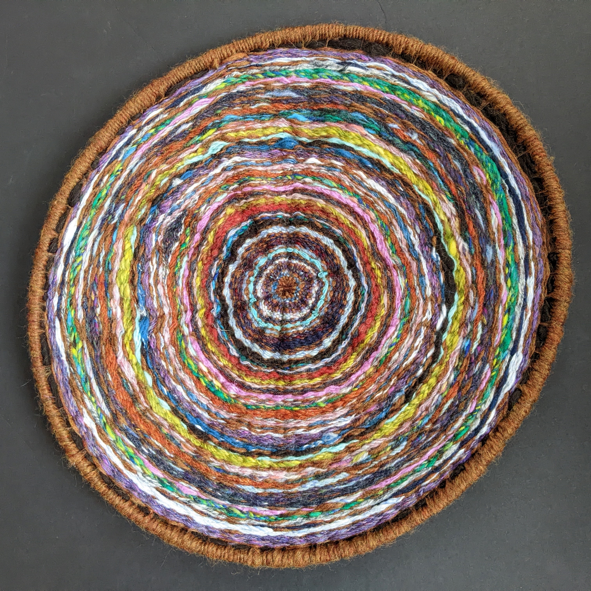 This piece of textile art shows yarn in a variety of colors woven in a circular shape. The rings of color mimic the rings on a tree.