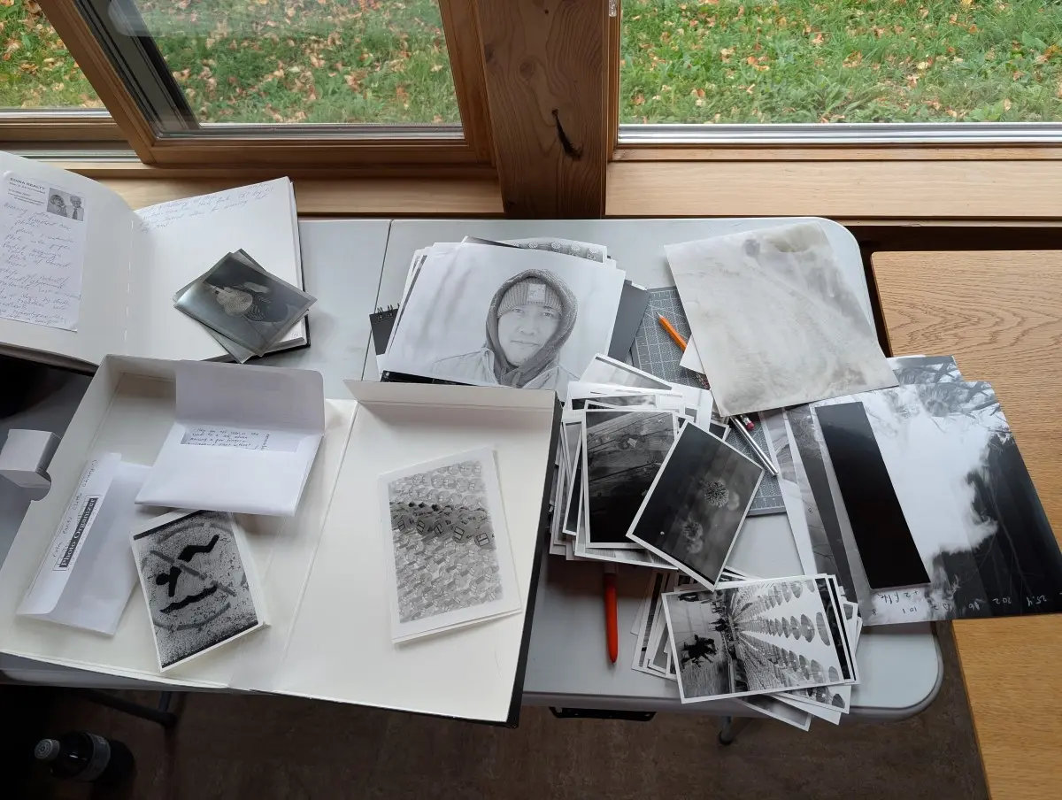 Several piles of photos rest on a folding table, along with a sketchbook, pen, pencil, and some envelopes.