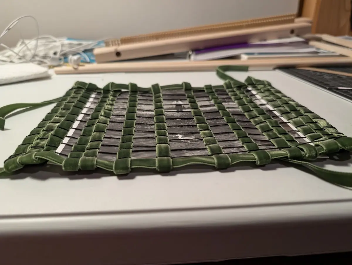 Sliced up photo strips that show a man looking at a heron are woven against a mossy green velvet ribbon. The black crochet cotton that was used as a warp thread makes the ribbon look like cobblestones. Weaving supplies are seen in the background.