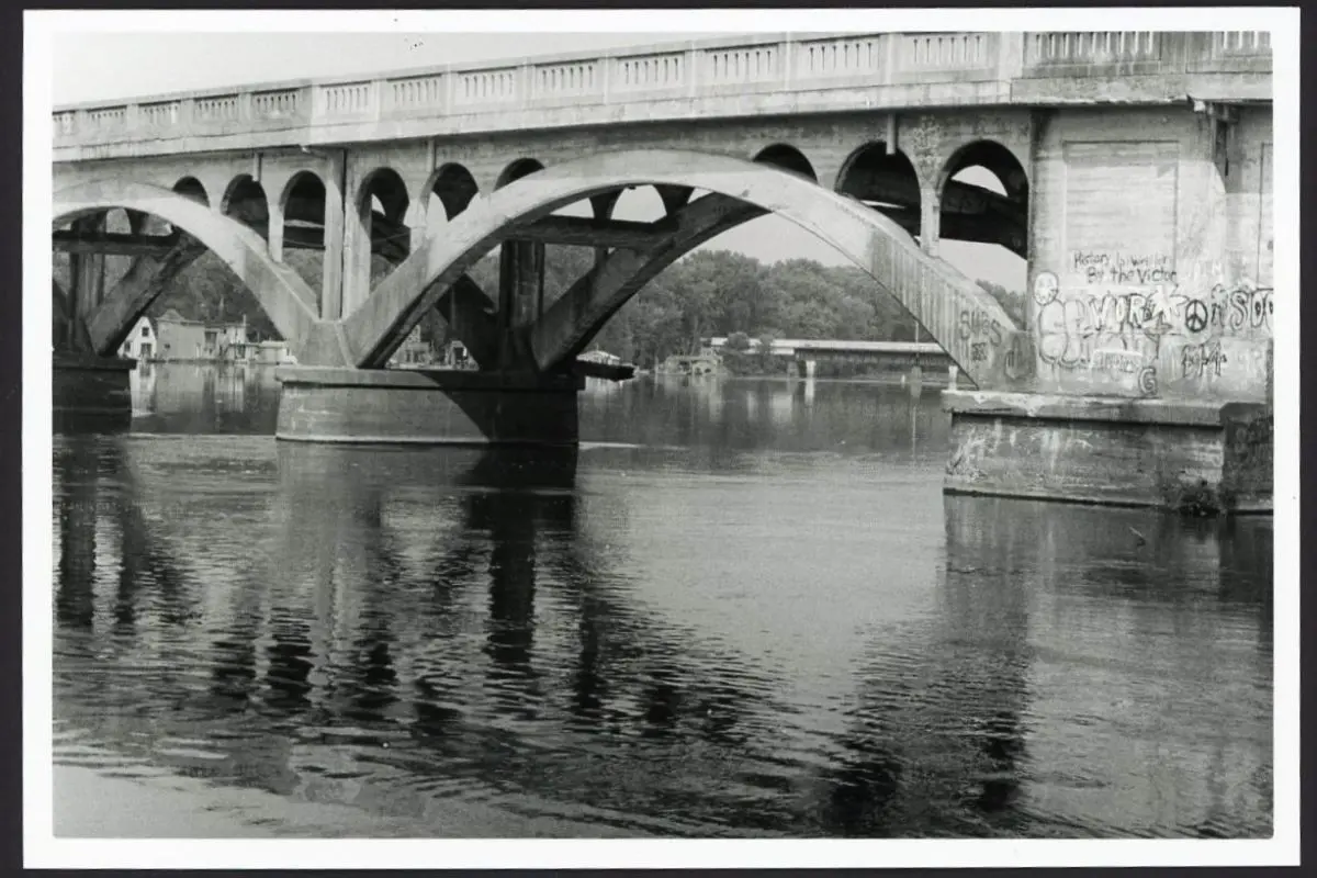 This black and white photo shows a bridge spanning the Mississippi River near Winona Minnesota. There is graffiti on the bridge.