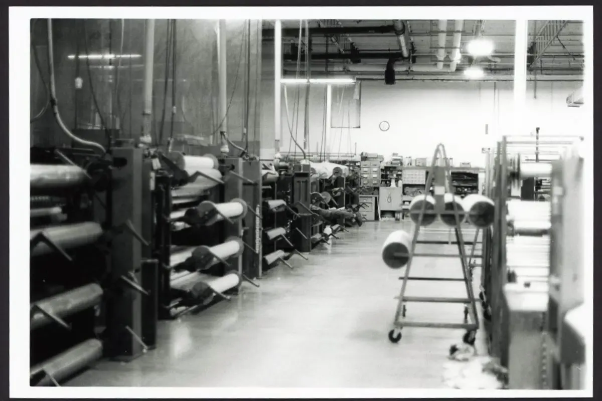 This black and white photo shows the inside of a factory. A man's legs can be seen sticking out from a machine.